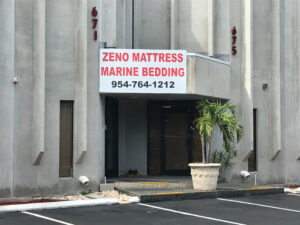 Image of Zeno Mattress storefront. Concrete building and black pavement parking.